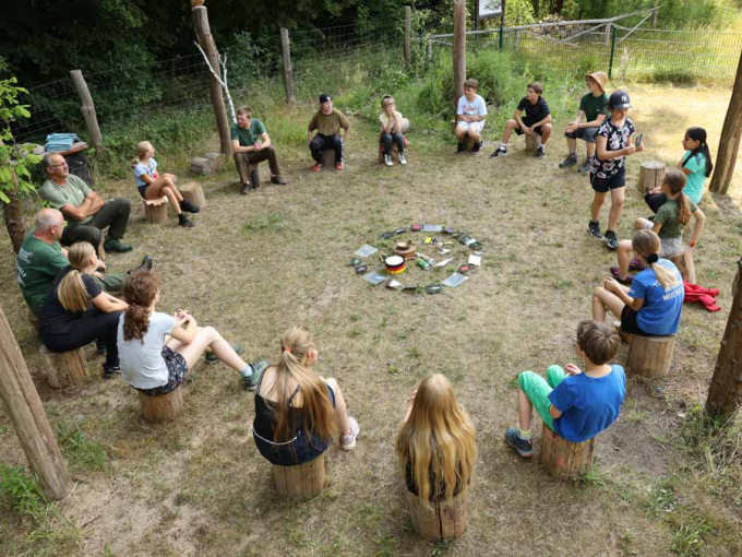 Kinder und Betreuer sitzen im Kreis auf kleinen Holzstämmen
